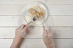 A girl with a knife and fork, cuts a piece of apple strudel with ice cream on white wooden table