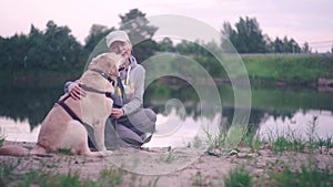 Girl with a Labrador retriever dog are sitting on sand near the water, laughing and kissing