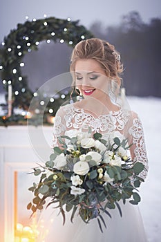 Girl in a lace wedding dress near the fireplace with Christmas decor on the background of snow-covered lake and the night sky.