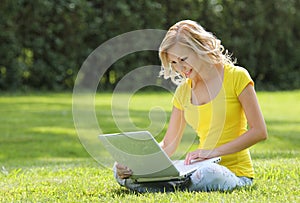 Girl with laptop. Blonde beautiful young woman with notebook sitting on the grass. Outdoor. Sunny day