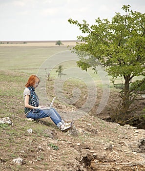 Girl with laptop at rock near lake and tree.