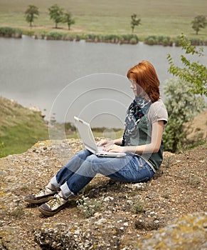 Girl with laptop at rock near lake and tree.