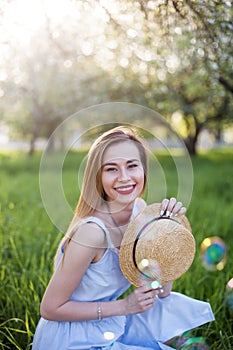 Girl laughs and holds a straw hat, red lips, blue sarafan. Happy girl rejoices in the summer, the sun and soap bubbles