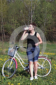 The girl leans on a parked bike. Rest on the spring cycle. The girl drinks water from a bottle