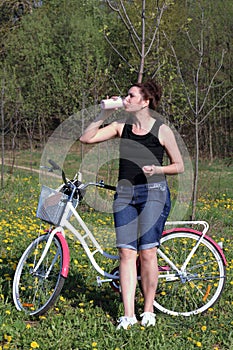 The girl leans on a parked bike. Rest on the spring cycle. The girl drinks water from a bottle