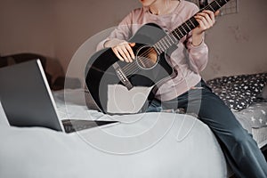 Girl learning to play guitar with the help of online learning at home. Woman sitting on bed with laptop and black guitar