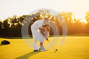 A girl learns to play golf, a man helps her by directing her stick before hitting