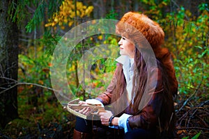 Girl in a leather jacket, a big red fox fur hat and with a crossbow in the forest in autumn. A female model poses as a