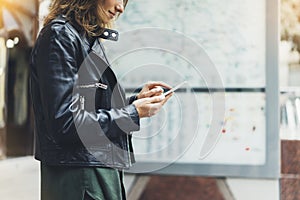 Girl in leather jacket holding smart phone on background map light box in sun atmospheric city, hipster using in female hands