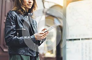 Girl in leather jacket holding smart phone on background map light box in sun atmospheric city, hipster using in female hands