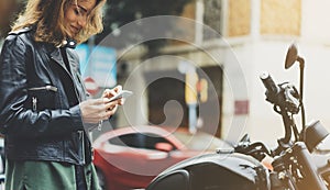 Girl in leather jacket holding smart phone on background motorcycle in sun atmospheric city, hipster using in female hands