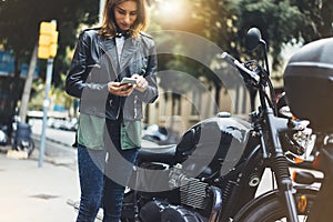 Girl in leather jacket holding smart phone on background motorcycle in sun flare atmospheric city, hipster using in female hands