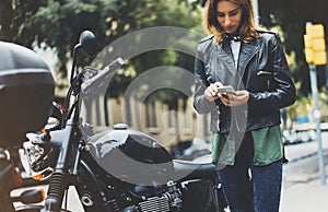 Girl in leather jacket holding smart phone on background motorcycle in sun flare atmospheric city, hipster using in female hands