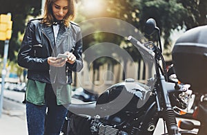 Girl in leather jacket holding smart phone on background motorcycle in sun flare atmospheric city, hipster using in female hands