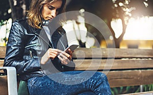 Girl in leather jacket holding smart phone on bench background sun atmospheric city, hipster using in female hands and texting