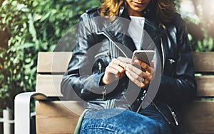 Girl in leather jacket holding smart phone on bench background sun atmospheric city, hipster using in female hands and texting