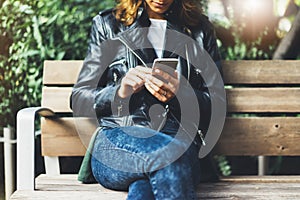 Girl in leather jacket holding smart phone on bench background sun atmospheric city, hipster using in female hands and texting