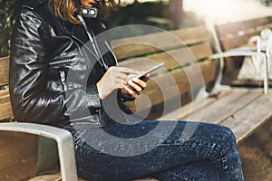 Girl in leather jacket holding smart phone on bench background sun atmospheric city, hipster using in female hands and texting