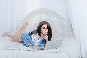 A girl lies on a bed with a laptop in a jeans jacket on a white background