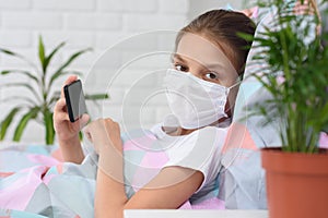 The girl lies on the bed in a medical mask with a mobile phone and looked into the frame