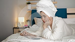 Girl reading book. Woman in bedroom with towel on her head and in white coat lies on stomach in bed and turns over page of book.