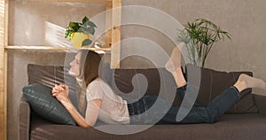 Girl lies on couch and swing ones feet. Young woman resting in living room during day.