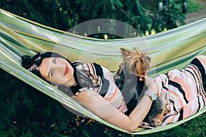 Girl lies in a hammock in nature with a dog in her arms and looks into the camera