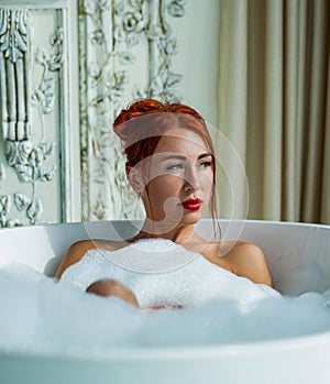 Girl lies in a large bath in foam. Beautiful woman taking bath with foam indoors. Beautiful young redhead girl takes
