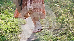 A girl in a light dress with a straw hat in her hand is walking through a forest clearing. Close-up of the legs. Slow motion.