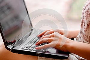 Girl in a light knitted sweater sits near the window and holds a laptop. Female hands print on a notebook, remote work at home