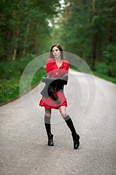 A girl in a light red airy dress, a leather jacket in a rockstyle on an empty road in a summer forest