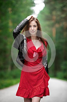 A girl in a light red airy dress, a leather jacket in a rockstyle on an empty road in a summer forest