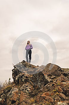 A girl in a lilac jacket looks out into the distance on a mountain, a view of the mountains and an autumnal forest by an overcast