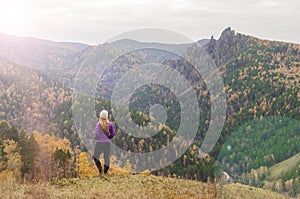 A girl in a lilac jacket looks out into the distance on a mountain, a view of the mountains and an autumnal forest by an overcast