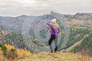 A girl in a lilac jacket looks out into the distance on a mountain, a view of the mountains and an autumnal forest by an overcast