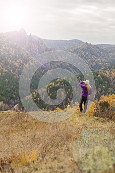 A girl in a lilac jacket looks out into the distance on a mountain, a view of the mountains and an autumnal forest by an overcast