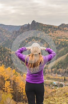 A girl in a lilac jacket looks out into the distance on a mountain, a view of the mountains and an autumnal forest by an overcast