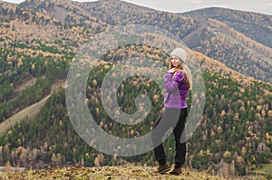 A girl in a lilac jacket looks out into the distance on a mountain, a view of the mountains and an autumnal forest by an overcast