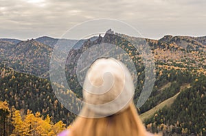 A girl in a lilac jacket looks out into the distance on a mountain, a view of the mountains and an autumnal forest by an overcast