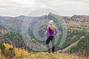 A girl in a lilac jacket looks out into the distance on a mountain, a view of the mountains and an autumnal forest by an overcast