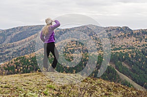 A girl in a lilac jacket looks out into the distance on a mountain, a view of the mountains and an autumnal forest by an overcast