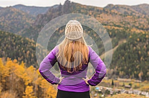 A girl in a lilac jacket looks out into the distance on a mountain, a view of the mountains and an autumnal forest by an overcast