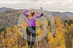 A girl in a lilac jacket makes a salfi on a mountain, a view of the mountains and an autumnal forest by a cloudy day