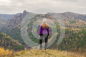 A girl in a lilac jacket standing on a mountain, a view of the mountains and an autumn forest