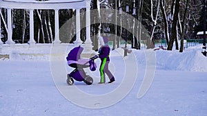 A girl in a lilac jumpsuit rolls a baby stroller in a city park against the background of a fountain and snow-covered trees in win