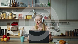 Girl listening music headphones sitting at kitchen with laptop. Woman relaxing.