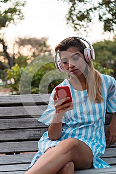 Girl listening to music on a headset and playing on a smartphone. Woman sitting on park bench listening to music