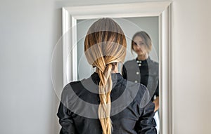 A girl with long blond hair braided in a braid is standing indoors opposite the mirror