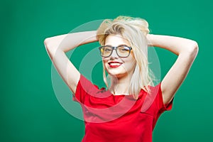 Girl with Long Blond Hair, Eyeglasses and Charming Smile Wearing Red Top is Posing on Green Background in Studio.