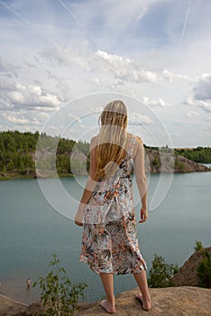 A girl with long blond hair in a light dress and bare feet stands on a cliff above the river